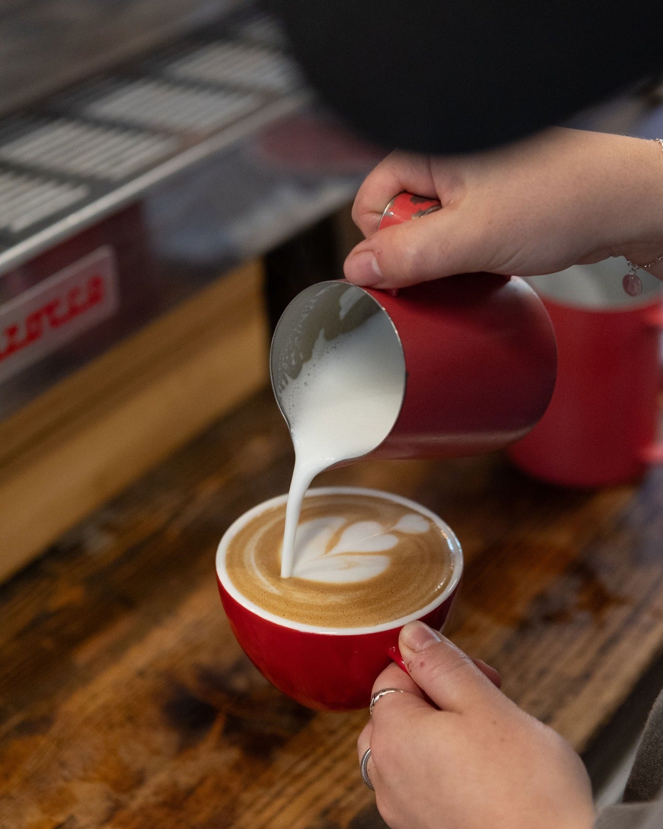 Person pouring milk into a red cup of coffee on a wooden surface.